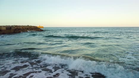 Waves-gently-crash-on-Costa-Garraf-beach-with-a-distant-view-of-a-cement-factory-in-Barcelona
