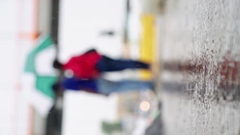 Vertical-video,-rainfall-on-puddle-in-concrete-with-people-out-of-focus