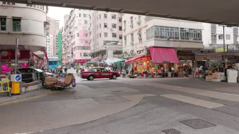 Die-Einheimische-Asiatische-Bevölkerung-Erledigt-Lebensmitteleinkäufe-Auf-Dem-Alten-Markt-Von-Kwa-Wan,-Während-Rote-Taxis-Und-Autos-Langsam-über-Die-Kreuzung-Unter-Der-Hochstraße-In-Hongkong-Fahren