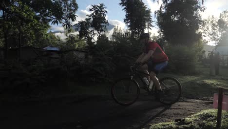 Marathon-athlete-cyclist-on-a-dirt-road-with-mountain-bike-riding-very-fast-towards-a-corner-and-mud-puddle-in-jungle-forest-Tanzania-with-lens-flare---slowmotion