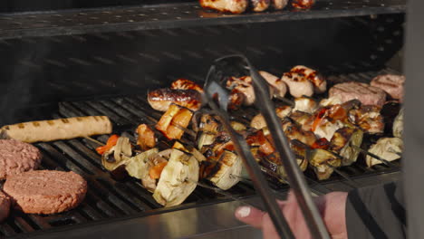 Chef-Hand-Turning-Vegetable-Shish-Kebabs-on-BBQ-with-Beef-Hamburgers-and-Sausages