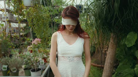 portrait-dolly-in-cinematic-video-of-a-Young-woman-in-white-dress-and-blindfolded-looking-at-camera-in-greenhouse-of-plants