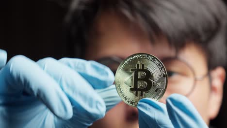 Asian-man-in-gloves-carefully-examines-a-gleaming-golden-bitcoin-cryptocurrency