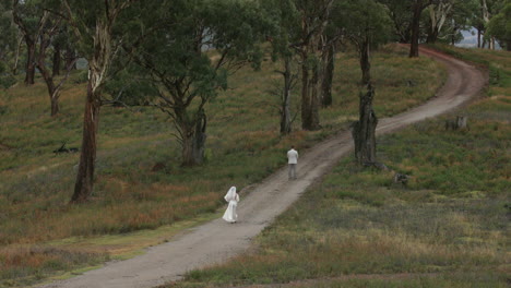 A-bride-in-her-white-dress-and-veil-walks-towards-the-groom-who-is-on-his-back-waiting-for-her-in-the-middle-of-a-road