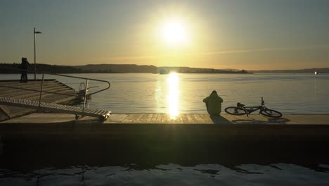 Person-Mit-Fahrradsilhouette,-Die-Vom-Hafen-Aus-Den-Sonnenuntergang-über-Dem-Oslofjord-Beobachtet