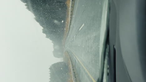 POV-car-driving-during-misty-heavy-snow-along-road-next-to-forest-with-wipers-cleaning-windshield