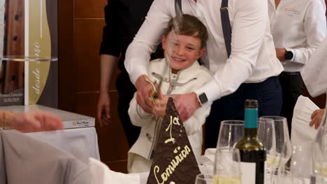 Little-Boy-Breaking-A-Tower-of-Chocolate-With-a-Sword-With-The-Help-of-His-Father-Over-His-Shoulder-During-A-Primera-Communion