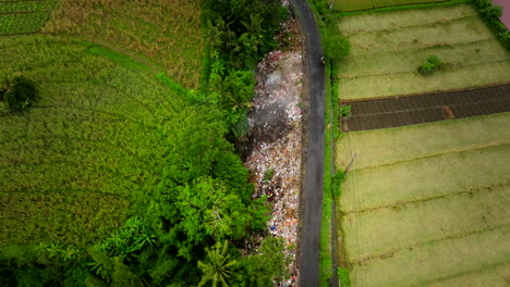 Heaps-of-trash-dumped-next-to-country-road-in-between-lush-farm-lands,-drone