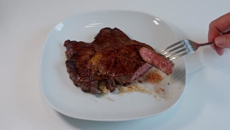 Closeup-of-a-fork-lifting-a-juicy-cut-piece-of-medium-cooked-entrecote-from-a-white-plate,-ready-to-eat