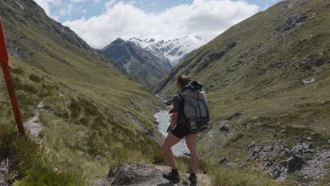 Young-caucasian-woman-walking-and-looking-around-in-a-valley-in-between-mountains-on-a-sunny-summer-day-in-Rees-Dart-track,-New-Zealand