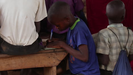Young-boy-in-a-blue-shirt-focuses-on-writing-at-a-wooden-desk-surrounded-by-other-children-in-Uganda