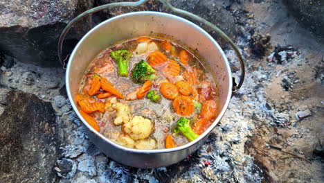 Campfire-cooking,-a-delicious-camp-oven-stew-with-vegetables-simmering-over-embers-in-a-fire-pit
