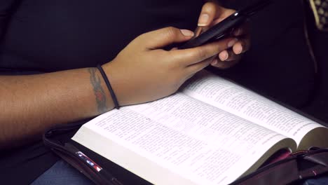 African-American-Black-Millennial-with-tattoos-using-cellphone-technology-while-reading,-studying-bible,-Christianity,-The-word-of-God